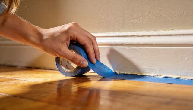 Applying blue painter's tape along baseboard for trim painting on hardwood floor
