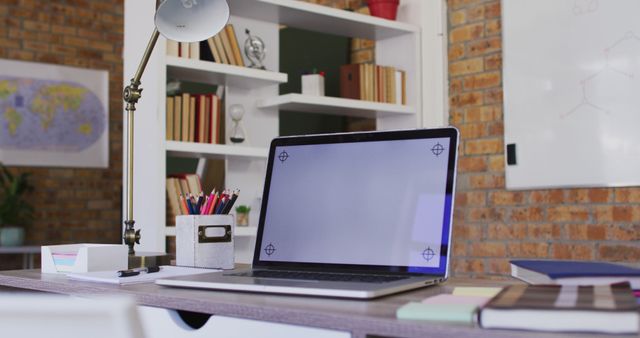 Modern workspace showcasing a laptop on a desk surrounded by office supplies such as books, pencils, and a lamp. Ideal for illustrating home office setup, remote work environment, or educational space. Suitable for articles, blog posts, or advertisements about productivity, work from home, or office organization.