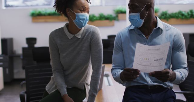 Colleagues engaging in conversation while examining a document in a modern office meeting room, both wearing masks for safety. Useful for themes related to workplace safety during the pandemic, professional collaboration, corporate environments, and health protocols in business settings.