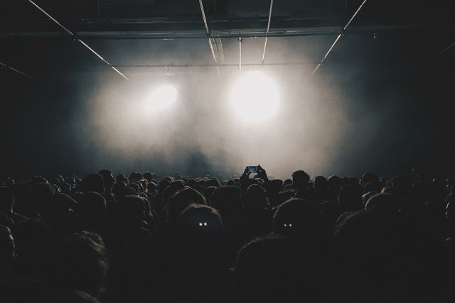 Silhouettes of concertgoers are facing a brightly lit stage at a live music event. Perfect for illustrating the atmosphere of a concert, event marketing, or articles about live performances.