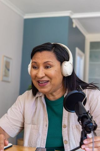Mature woman podcasting from home wearing white headphones and speaking into microphone