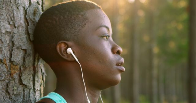 Young woman with short hair leaning against tree in forest, listening to music through earbuds. Capturing a moment of solitude and contemplation during a sunny day in nature. Ideal for concepts related to relaxation, mental health, and outdoor activities.