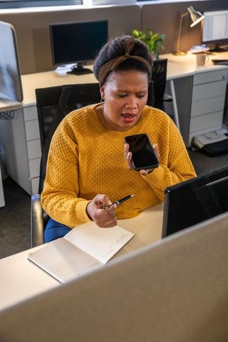 African american woman speaking into smartphone, pointing at notes while working at desk