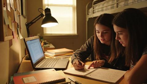 Two teen girls studying late at desk with laptop lamp on collaborating on homework