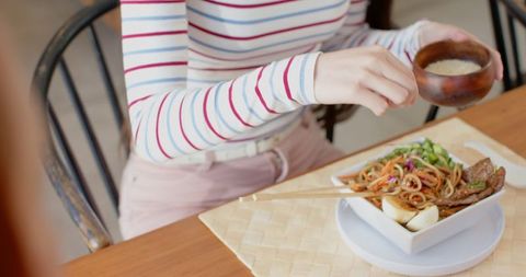 Woman adding seasoning to asian noodles with chopsticks on table