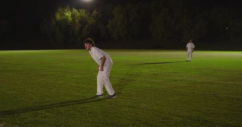 Night cricket players under floodlights preparing for delivery in white kits and shadows