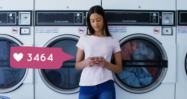 Close up of a biracial woman typing on her mobile phone while waiting for her laundry to finish in laundry shop. Pink digital image of a message bubble with a heart icon and a number count up.