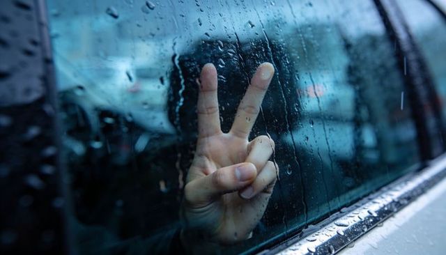 Peace sign hand against rain-soaked car window with moody urban reflection, chrome sill