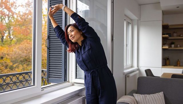 Stretching by open window in cozy autumn apartment wearing navy jumpsuit