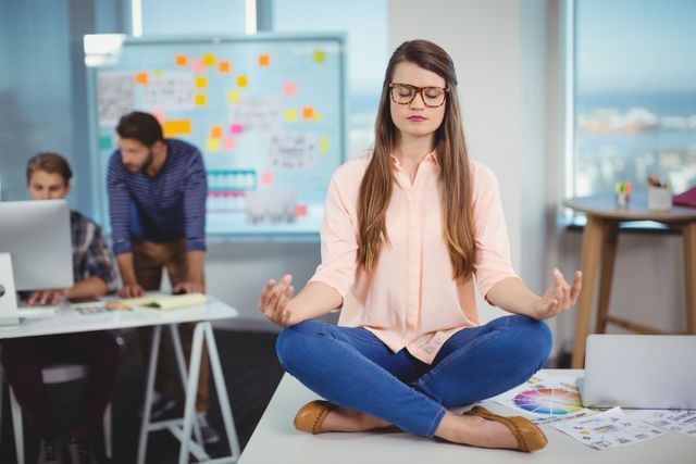 Female graphic designer sitting cross-legged on office desk, meditating with eyes closed. Colleagues working in background. Useful for illustrating workplace wellness, stress management, mindfulness practices, and promoting a balanced work environment.