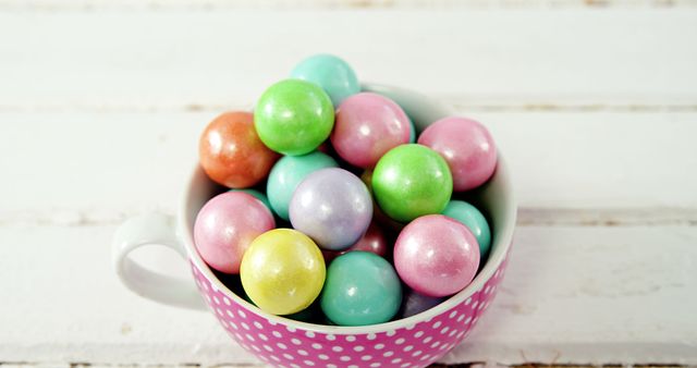 Pastel-colored candy gumballs are filling a pink polka dot cup on a weathered white wooden table. This vibrant and playful image can be used in advertisements for candy brands, party planning materials, or creative dessert recipes. It also adds a pop of color and a sense of fun to blogs and social media posts.