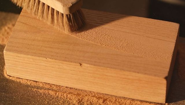 Stiff-bristle brush brushing sawdust from planed wood block on woodworking bench under warm light