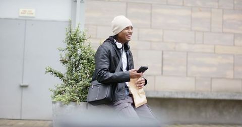 Young man laughing while checking smartphone outdoors with headphones and messenger bag