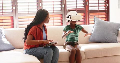 African mother guiding child using vr headset on sofa for home learning and play