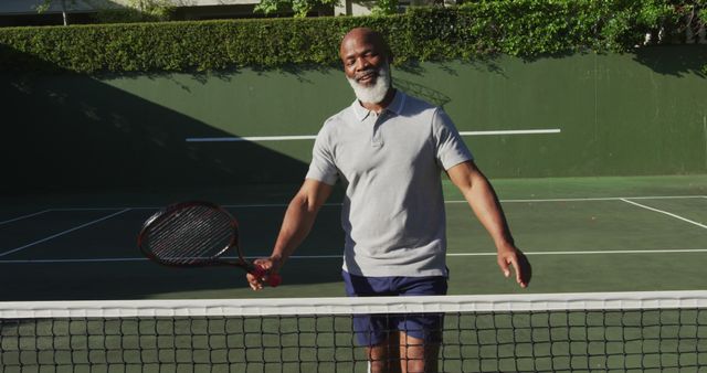 Senior man standing on an outdoor tennis court smiling while holding a tennis racket. Ideal for use in promoting healthy lifestyles, active aging, sports participation, and recreational tennis activities. Also great for advertisements or articles aimed at seniors, highlighting leisure activities and fitness.