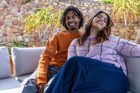Diverse couple smiling and relaxing on outdoor sofa with succulents, stone wall backdrop