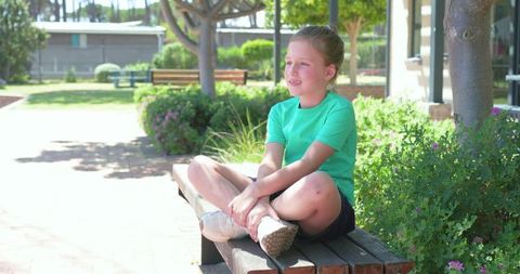 Smiling young girl sitting cross-legged on wooden bench in sunny park wearing green shirt