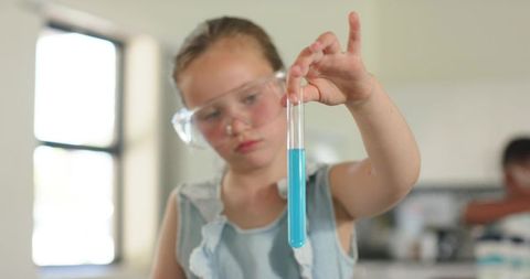 Girl inspecting blue liquid in test tube while wearing safety goggles in classroom