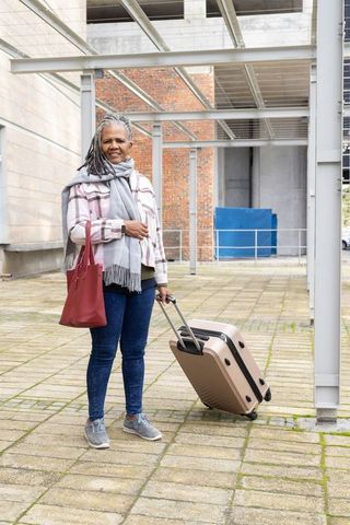 Senior african american woman standing with suitcase and red tote in urban courtyard