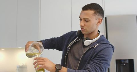 Young man pouring green smoothie from blender into tall glass in modern kitchen