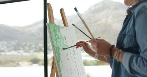 Mature female painting on canvas at easel with mountain view through large window