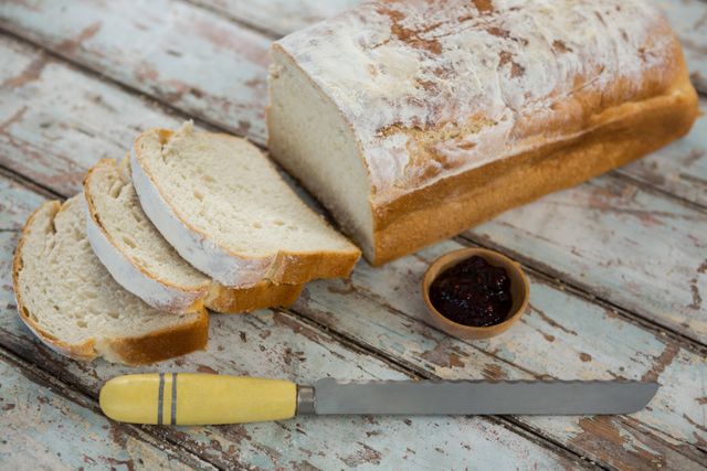 Freshly sliced bread with a small bowl of jam and a knife on a rustic wooden table. Ideal for use in food blogs, bakery advertisements, breakfast recipes, and culinary websites. Perfect for promoting homemade and artisan bread products.