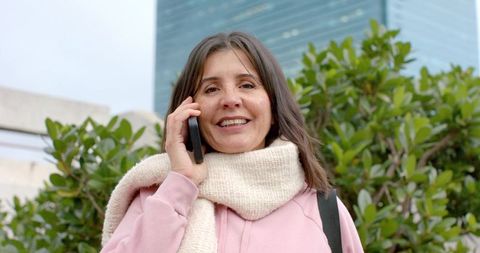 Mature woman smiling and talking on smartphone on rooftop terrace in pink jacket
