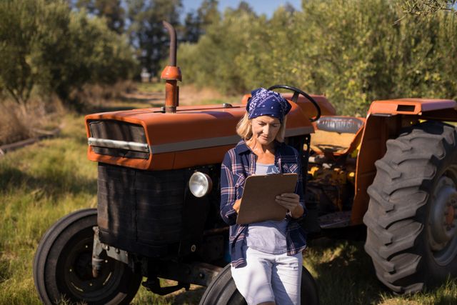 Woman leaning on tractor while writing on clipboard in olive farm