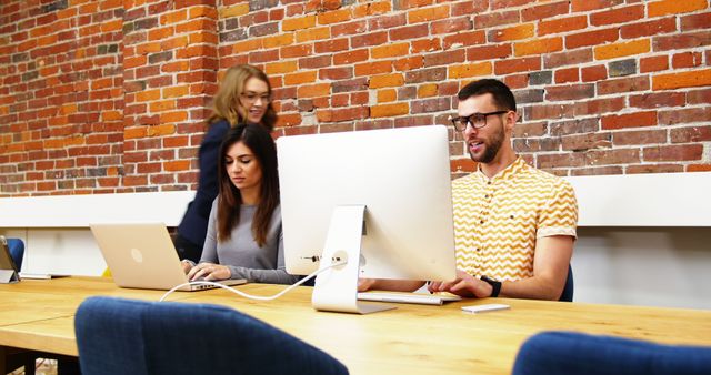 A diverse group of young professionals is working diligently in a modern office space with exposed brick walls, with copy space. The setting suggests a collaborative and trendy workplace environment.