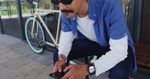 Man wearing sunglasses and blue casual shirt, sitting outside with a vintage bicycle in the background, checking his smartphone. Can be used to depict modern lifestyle, technology use, cycling enthusiasts, casual outfits, or outdoor relaxation.