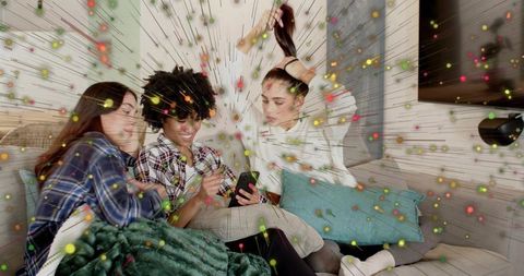 Friends sharing smartphone on couch surrounded by colorful digital burst and cozy blankets