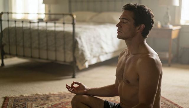 Shirtless man meditating cross-legged on rug beside metal bed in sunlit bedroom