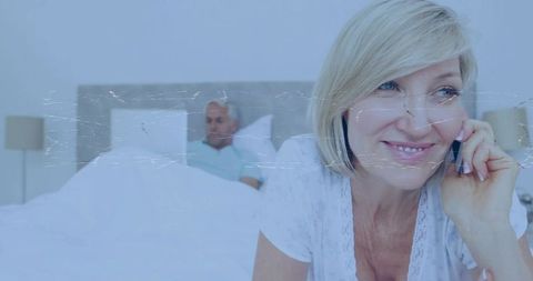 Mature woman talking on smartphone in bed while partner resting nearby, intimate morning scene