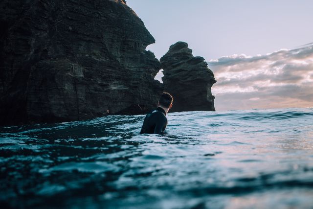 Person swimming near rocky cliffs during dusk in open ocean. Horizon shows slight cloud formation and serene environment. Suitable for blogs, travel advertisements, nature magazines, inspirational posters, or websites focused on adventure and outdoor activities.