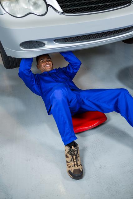 Mechanic in blue coveralls smiling while working under a car in a garage. Ideal for use in automotive service advertisements, repair shop promotions, and articles about car maintenance or professional mechanics.