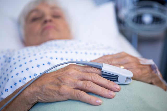 Elderly patient resting in hospital bed with pulse oximeter on hand, monitoring vital signs. Useful for healthcare, medical technology, patient care, and hospital-related content. Can be used in articles, blogs, and educational materials about elderly care, medical monitoring, and hospital equipment.