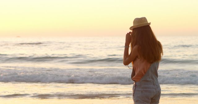 Young woman wearing a hat taking photos by the ocean during a beautiful sunset. Ideal for themes related to travel, vacations, summer getaways, and relaxation. Useful for brochures, travel articles, advertising campaigns, and social media posts focusing on outdoor and leisure activities by the sea.