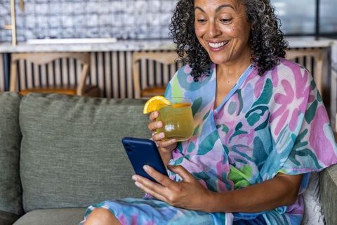 Middle-aged african american woman lounging on sofa with citrus cocktail and smartphone