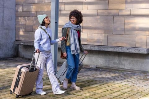Two diverse women walking through urban plaza pulling rose-gold and grey rolling luggage