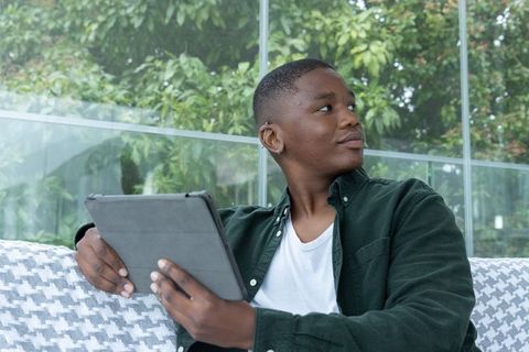 African american man sitting on sofa holding tablet, glancing toward window in sunroom