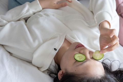 Asian woman relaxing with cucumber eye slices on bed in white zip-up pullover