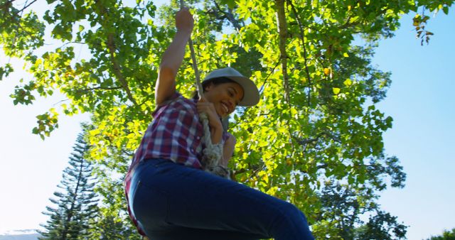 Happy biracial woman in cap hanging on rope and laughing in sunny park, copy space. Free time, lifestyle, relaxation and city break, unaltered.