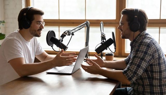 Two male podcast hosts recording conversational interview with laptop, microphones, coffee