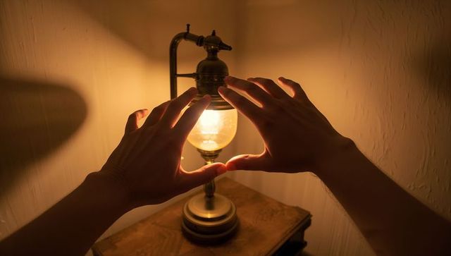 Hands cupping warm glowing bulb beside vintage brass lamp on wooden nightstand