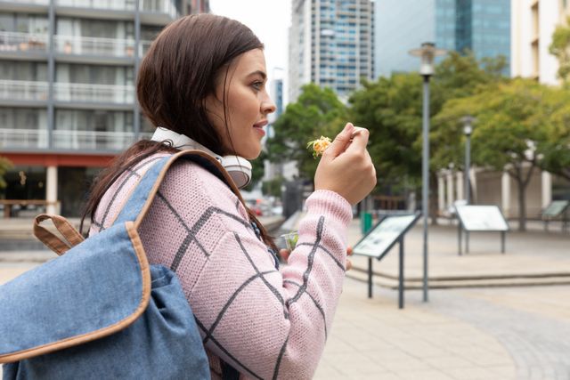 Curvy woman enjoying a healthy takeaway salad while standing in a city street during the day. She is wearing headphones and a rucksack, with modern buildings in the background. Ideal for use in urban lifestyle, healthy eating, and outdoor activity themes.