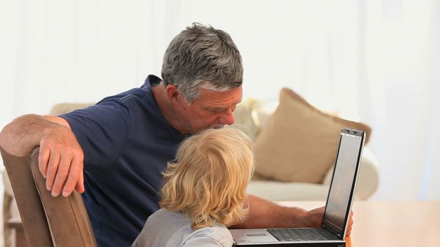 Elderly man and young boy are sitting together at home, engaged in looking at a laptop. Grandfather and grandson share a moment of connection and learning, showing the influence of technology even in family settings. Suitable for content highlighting family relationships, intergenerational learning, or technology use among different age groups.