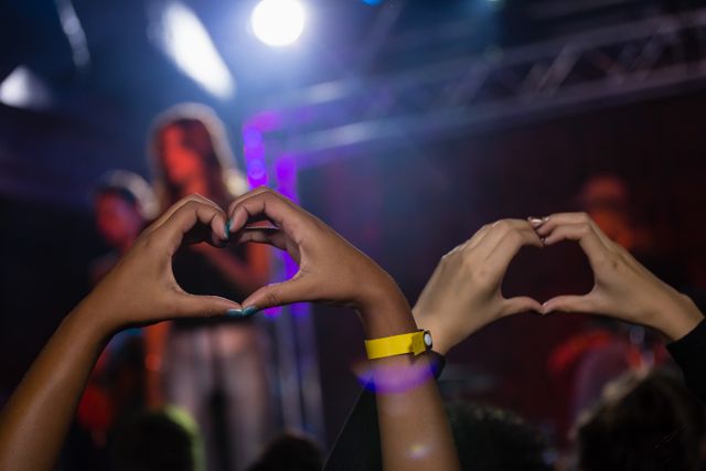 Hands of audience members forming heart shapes during a live concert in a nightclub. Ideal for use in promotions for music events, nightlife advertisements, and articles about live performances and fan culture.