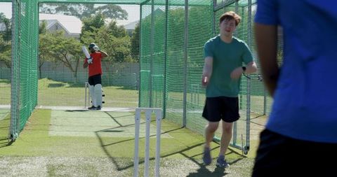 Cricket practice nets with batter waiting and teammate walking toward camera in sunny outdoor sessio