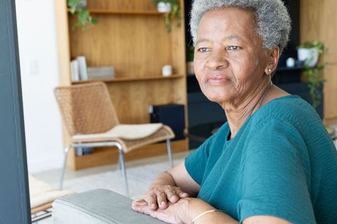Reflective senior african woman sitting on sofa armrest in sunlit modern living room