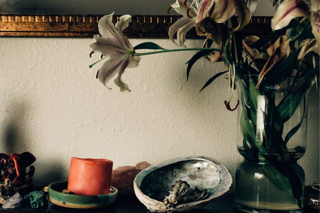 Interior showcasing a vase of lilies and a lit candle sitting on a table beside a decorative shell. Ideal for home decor inspiration, wellness blogs, or aromatherapy product promotions. Evokes peace and tranquility in residential spaces.